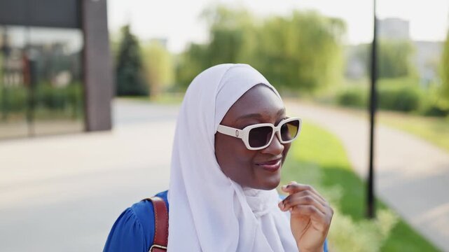 A young African American Muslim woman wearing a white hijab and sunglasses smiles coyly while adjusting them outdoors