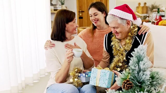 Smiling family celebrating Christmas at home with gifts by a decorated tree. An older man in a Santa hat and tinsel shares a warm moment with two women