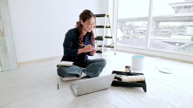 Young woman sits on the floor of a bright apartment, planning a DIY paint job with a laptop, roller tray, bucket, and ladder around her during home renovation