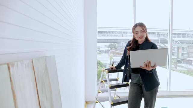 Interior designer with laptop inspecting a bright room under renovation, with ladder and panels by large windows overlooking an urban cityscape.