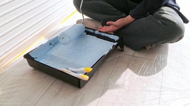 Person kneels on a protected floor, loading a roller with light blue paint in a tray, prepping an interior wall taped for clean edges.