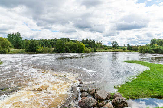 A scenic wide-angle view of a river weir with cascading water, white foam, and rocky foreground elements, featuring high water levels overflowing onto green grassy banks under a cloudy sky.