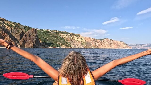 Carefree paddler stretching arms wide atop ocean waters near rocky coastline. Stationary shot features human back and tranquil sea backdrop.