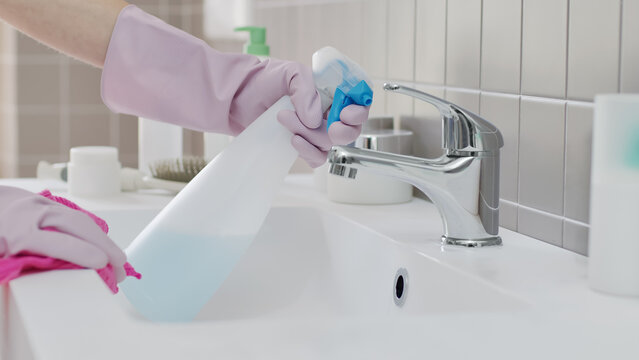 Close up of hands wearing rubber gloves spraying detergent and wiping chrome faucet with cloth