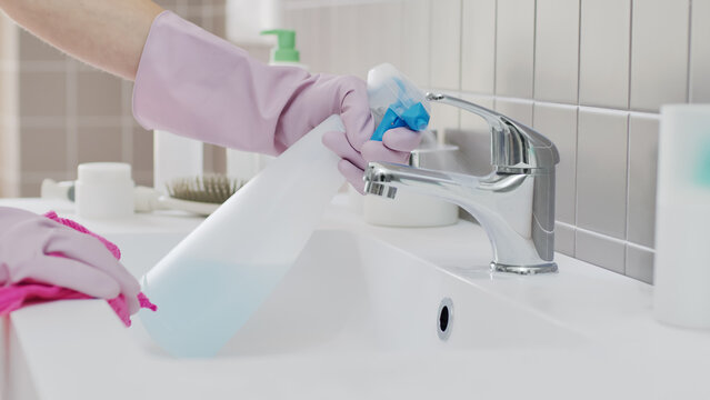 Close up of hands wearing rubber gloves spraying detergent and wiping chrome faucet with cloth