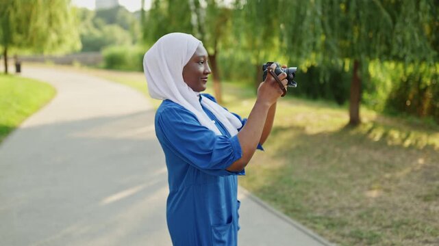 An African American Muslim woman wearing a hijab and blue dress captures moments with her camera in a sunlit park setting