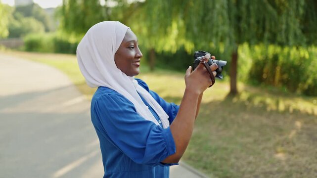 A smiling African American Muslim woman wearing a white hijab and blue dress holds a camera and takes pictures in a park