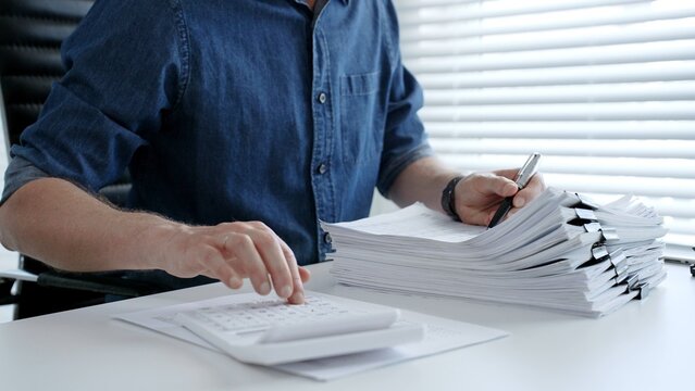 Individual working diligently on financial documents, calculating expenses and reviewing reports at an office desk, managing a large stack of paperwork and invoices