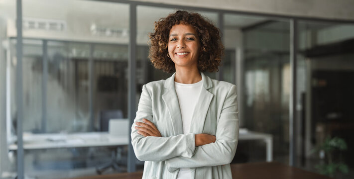 Portrait of confident latin business woman with arms crossed, smiling in modern glass office. Stylish businesswoman poses with assurance near open workspace. Banner, copy space.