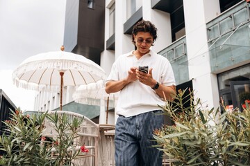 Obraz premium Young man in casual white shirt and striped pants using smartphone outdoors on a balcony with plants and an umbrella in a modern urban setting