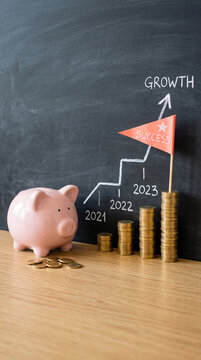 Pink piggy bank and stacked coins on a wooden surface in front of a chalkboard displaying a growth chart, symbolizing savings and financial success.