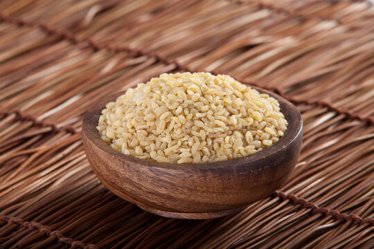 Raw bulgur wheat in wooden bowl on wicker mat surface.