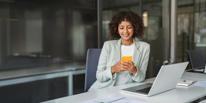 Smiling hispanic business woman specialist checking phone messages beside open laptop at office desk. Modern professional stays connected, managing daily tasks smoothly. Banner, copy space.