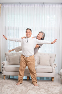 playful senior asian couple having fun together in living room at home,husband opening arms while a wife stands on a sofa behind,holding on his shoulders and laughing heartily