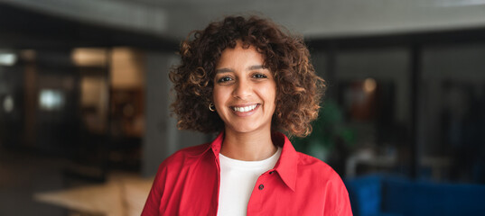 Fototapeta na wymiar Close up headshot portrait of smiling young hispanic woman with curly hair, casual style in office. Cheerful latina gazes at camera in relaxed, candid moment indoors. Banner, copy space.