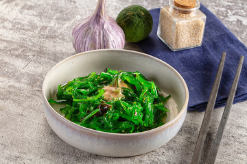 Fresh green seaweed salad in ceramic bowl with sesame dressing, garlic, lime and chopsticks on textured stone surface © Andrei Starostin