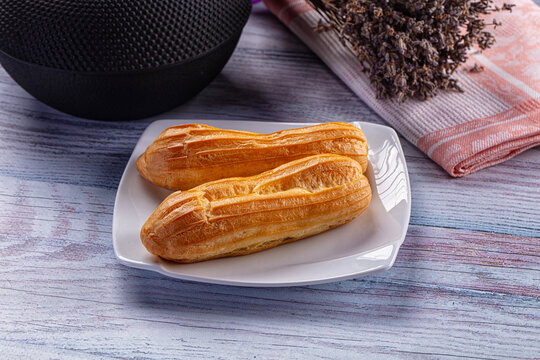 Two golden brown eclair pastries on white ceramic plate with textured black speaker and dried lavender on striped cloth background