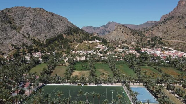 Aerial video zooming out from a green soccer field surrounded by palm trees at El Palmeral park in Orihuela, Spain. Stock footage for sports, travel, and geography projects.