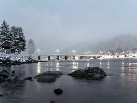 Snowy mountains and illuminated bridge over water in Artybash village near Teletskoye lake, Altai, Russia. Winter evening landscape with stones in river and copy space for travel banner design