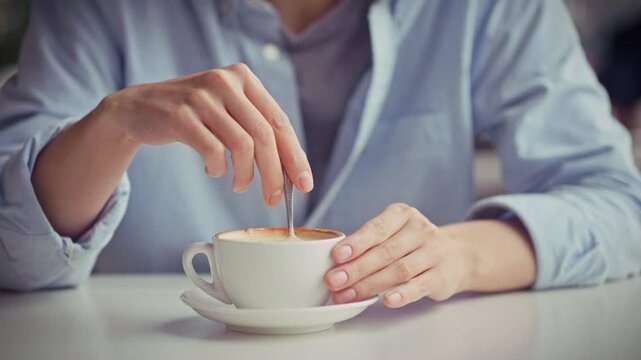 Stirring a cappuccino, the girl sits at a table in a coffee shop, mixing the foam in a ceramic cup with a spoon - theme of daily beverage preparation under natural daylight, quiet ambience