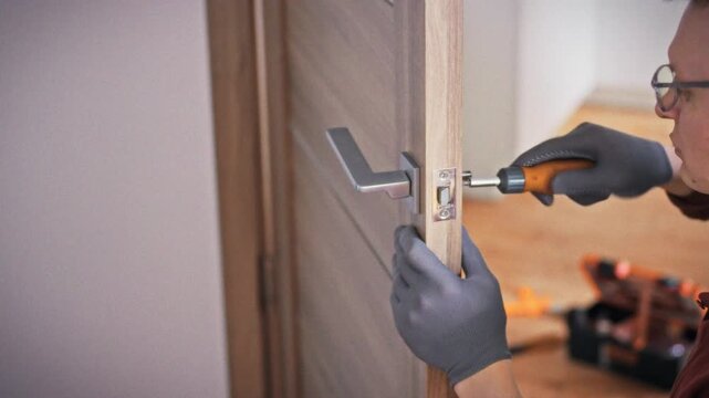 A close-up of an orange screwdriver in gloved hands working on a door lock mechanism on a grey brown wooden interior door. Concept of home door repair and lock maintenance