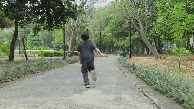 rear view of a young child running along a paved path through a tree lined park