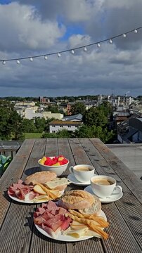 Delicious breakfast spread. with coffee cups, bread rolls, charcuterie, and fruit salad on a rustic wooden table overlooking the skyline of Rostock under cloudy sky.