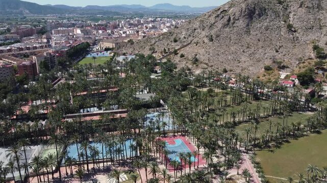 Aerial drone shot moving toward Sierra de Orihuela mountains, flying over a lush green palm grove, sports fields, and urban landscape in Alicante, Spain.