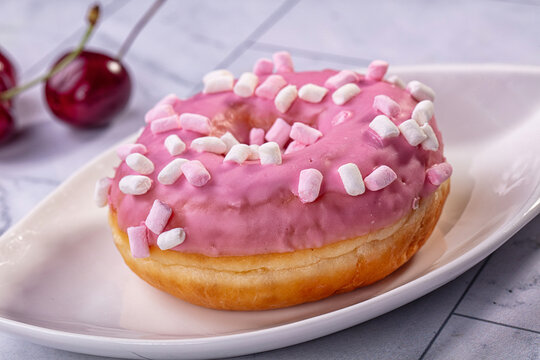 Pink frosted donut topped with mini marshmallows on white plate, fresh cherries in background, soft lighting, shallow depth of field
