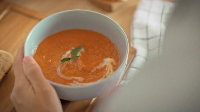 A housewife eats warm homemade tomato soup with croutons from a blue bowl using a wooden spoon. Pieces of fresh white bread on the wooden table beside. Concept of healthy homemade nutrition