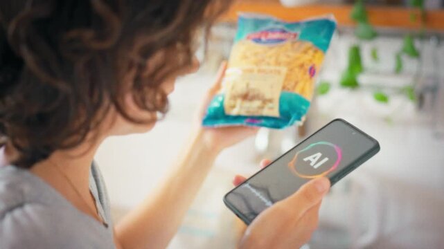 A housewife holds a pasta package and consults an AI assistant on her phone about the recipe. Idea of digital technology as a smart kitchen helper in everyday home life