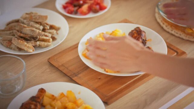 A housewife serves roasted meat and baked potatoes onto a plate from a full kitchen counter in daylight. Food preparation and serving process at home. Concept of homemade family meal