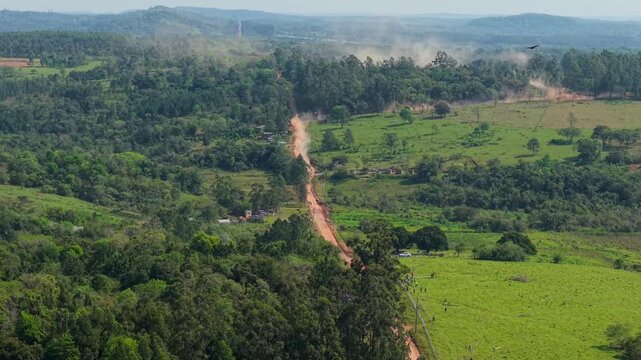 Long dirt rally road crossing green rural landscape in Nueva Alborada, Paraguay, aerial overview with rising dirt and dust from racing car
