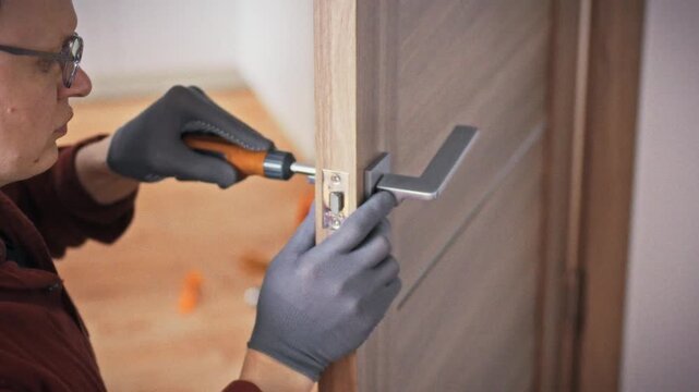 A profile view of a specialist in uniform carefully working on an interior door handle with an orange screwdriver against a background of an orange toolbox. Concept of DIY home door repair