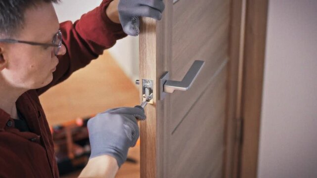 A technician in uniform and glasses methodically repairs an interior door lock using an orange screwdriver in a bright home setting. Concept of calling a specialist for domestic door repair