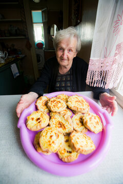 Elderly woman presenting a tray of baked pastries at a kitchen table by a window and looking at the camera