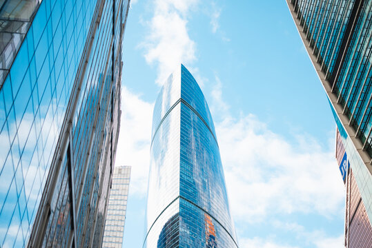 A tall, modern skyscraper stands between two glass buildings in an urban setting. The structure reflects the clear blue sky and scattered clouds, showcasing contemporary architectural designs