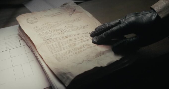 Leather-gloved hand places old secret documents onto a desktop in low light, evoking clandestine activity, undercover intelligence and vintage spy storytelling.