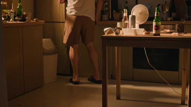 Caucasian middle aged man walking across kitchen, opening refrigerator, grabbing beer bottle, and resting it on forehead. Footage ideal for substance abuse campaigns and mental health awareness