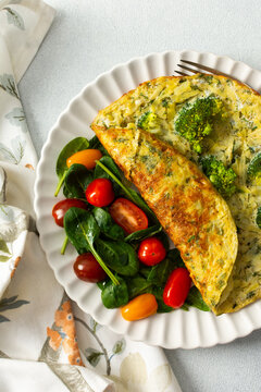 A potato omelette made with eggs and mixed greens is served on a white plate. Spinach, cherry tomatoes, and broccoli are arranged on the side for a healthy breakfast option