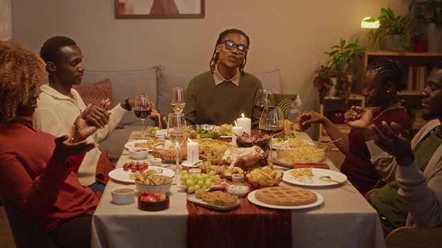 African American young adults holding hands, praying together at Thanksgiving dinner table before holiday feast. Footage fits festive holiday commercials, food advertising and community campaigns