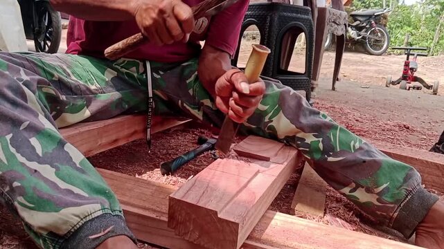 Man Carving Wood with Hammer and Chisel. 
A man carving and shaping wood with a hammer and chisel during a traditional woodworking activity in a rural outdoor setting.