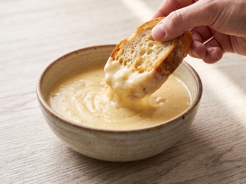 Hand dipping a slice of toasted crusty bread into a warm bowl of creamy cheese fondue soup