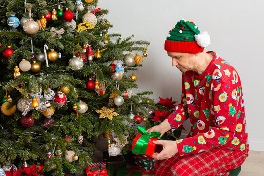 A 53 year old man sits near a decorated Christmas tree on Christmas Day. He is opening a gift while wearing a festive outfit and a holiday hat. Family and joy fill the space around him