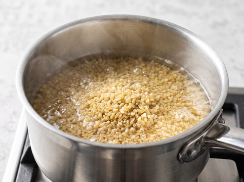 Raw bulgur wheat groats boiling in clean water inside a stainless steel saucepan