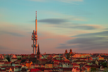 Obraz premium Žižkov Television Tower rising above Prague at sunset, with St. Procopius Church and red-roofed cityscape glowing in warm evening light, Czech Republic.