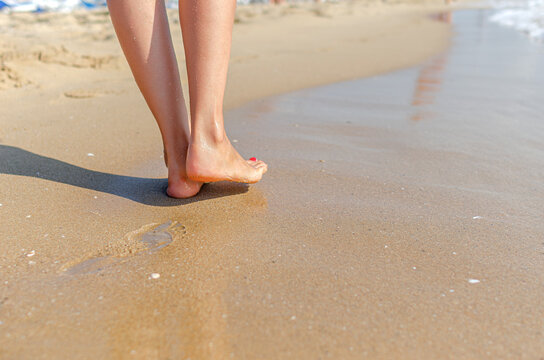 A woman walks barefoot along the wet sand at the edge of the ocean on a sunny day.