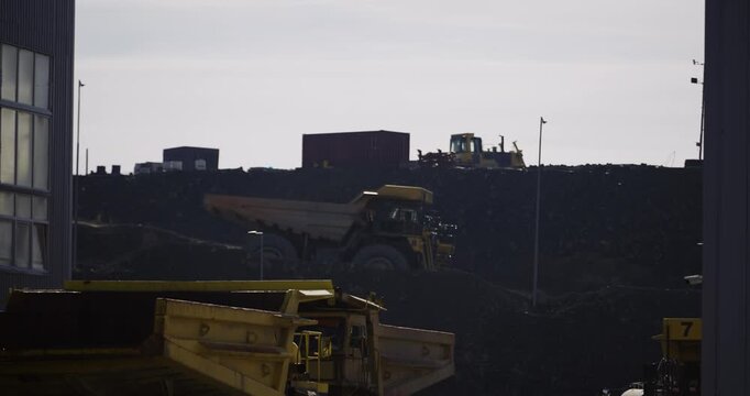A large yellow dump truck is parked next to a building. The truck is surrounded by other yellow trucks. The scene is industrial and the trucks are likely used for construction or waste management