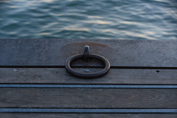 Obraz premium Close-up of a sturdy steel mooring ring on a wooden dock with calm water ripples in the background at Lake Garda. Concept of maritime equipment, boat mooring, nautical transport, pier construction, an
