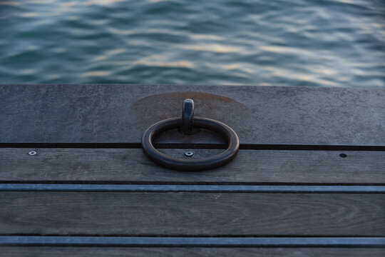Close-up of a sturdy steel mooring ring on a wooden dock with calm water ripples in the background at Lake Garda. Concept of maritime equipment, boat mooring, nautical transport, pier construction, an